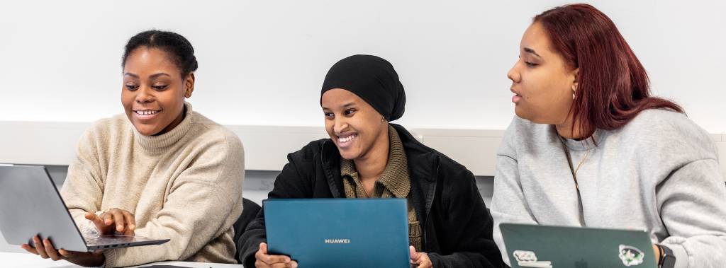 Tre kvinnelige studenter smiler og snakker sammen i klasserommet Fagskolen Diakonova