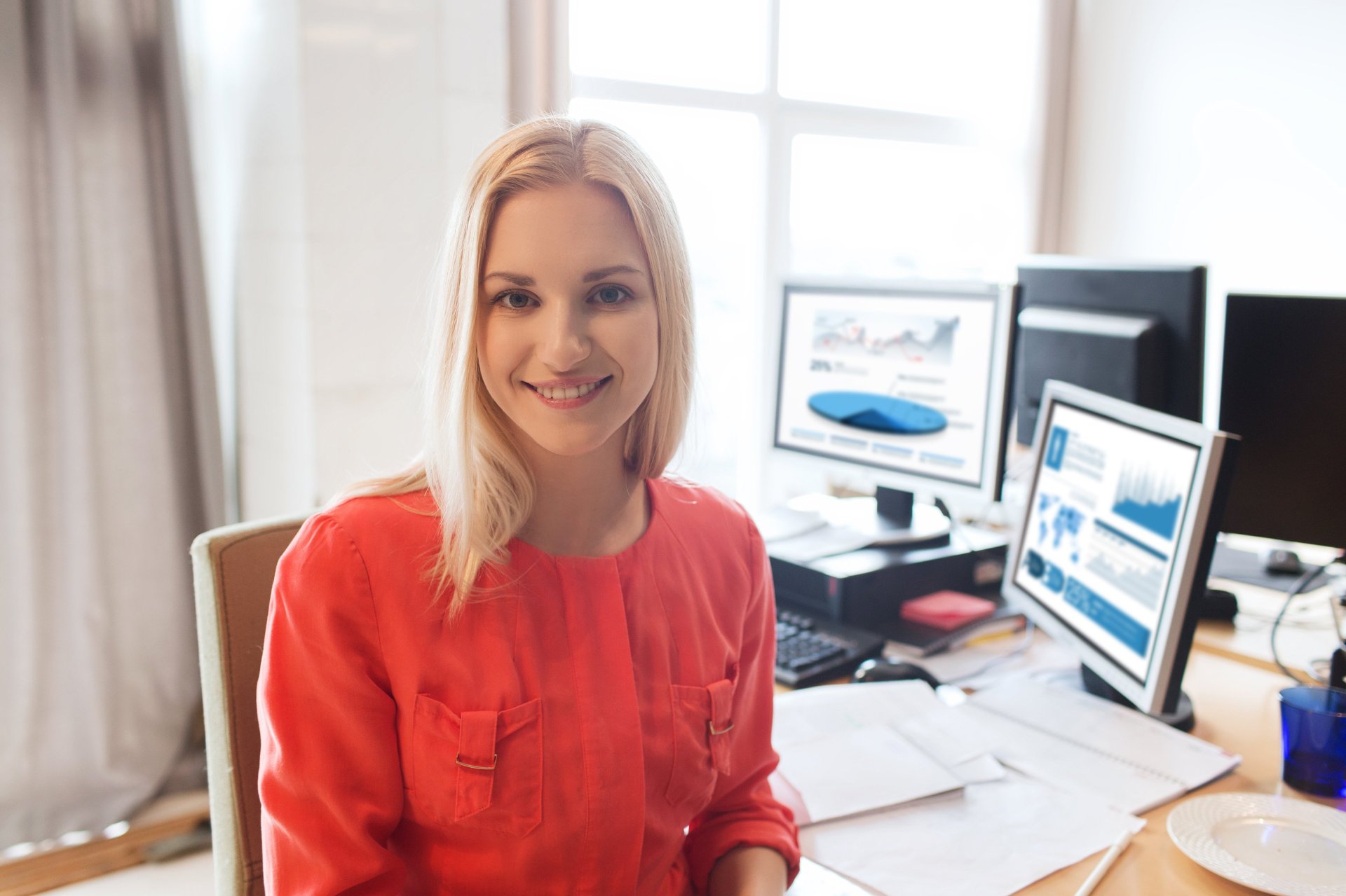 Kvinne som sitter ved en pult med skjermer og smiler og ser mot kamera