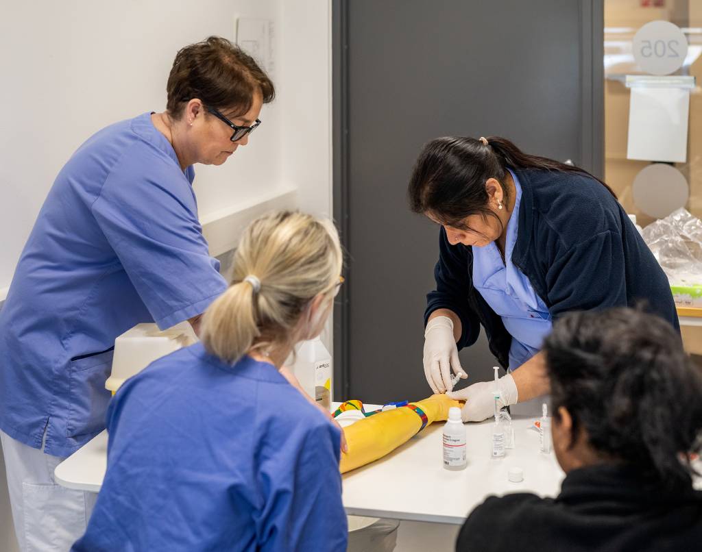 Fire helsearbeidere rundt et bord, tre ser på mens den fjerde stikker en sprøyte i en kunstig arm.