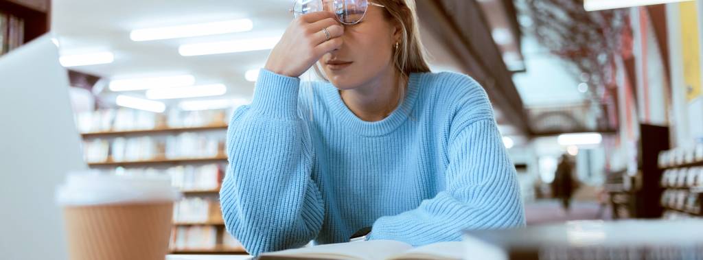 Jente sitter på bibliotek. Tar seg til hodet og ser sliten ut.