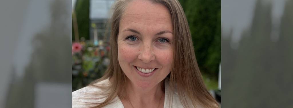 Kristin smiler til kamera foran en grønn hage med blomster i bakgrunnen