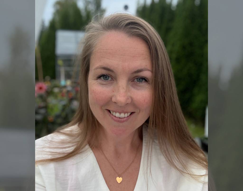 Kristin smiler til kamera foran en grønn hage med blomster i bakgrunnen
