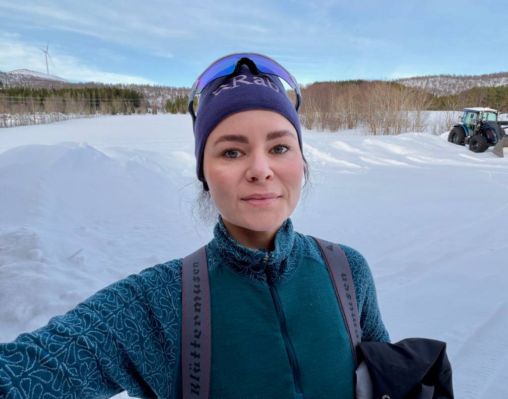 Sara står ute en vinterdag med skog og en traktor i bakgrunnen
