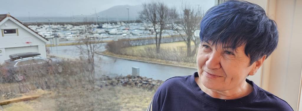 Ljilja sitter foran et vindu med en båthavn i bakgrunnen