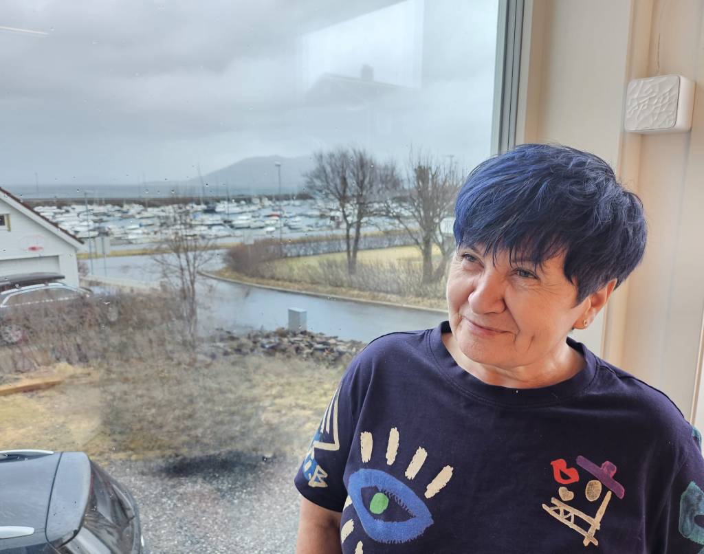 Ljilja sitter foran et vindu med en båthavn i bakgrunnen