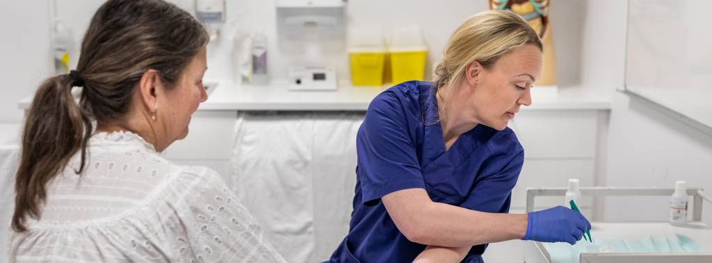 Kvinnelig helsefagarbeider steller sår hos kvinnelig pasient som sitter med ryggen til