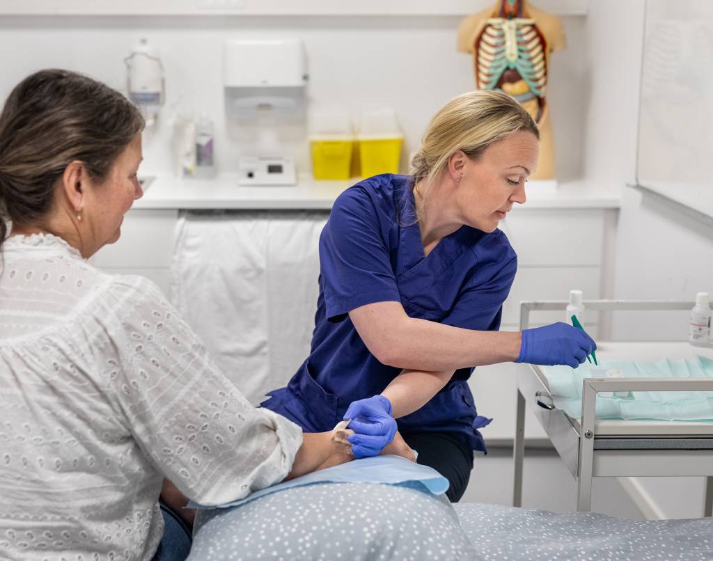 Kvinnelig helsefagarbeider steller sår hos kvinnelig pasient som sitter med ryggen til