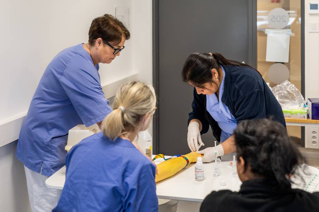 Fire helsearbeidere rundt et bord, tre ser på mens den fjerde stikker en sprøyte i en kunstig arm.!''