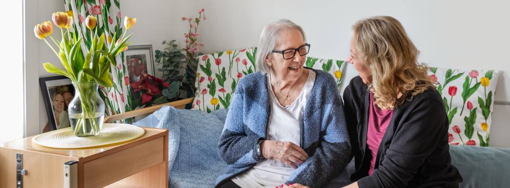 Mor og datter sitter sammen på en seng og smiler til hverandre. Fagskolen Diakonova