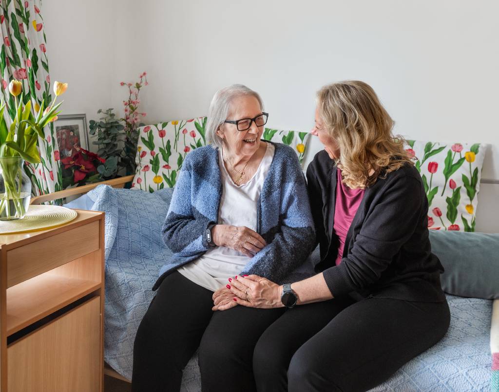 Mor og datter sitter sammen på en seng og smiler til hverandre. Fagskolen Diakonova